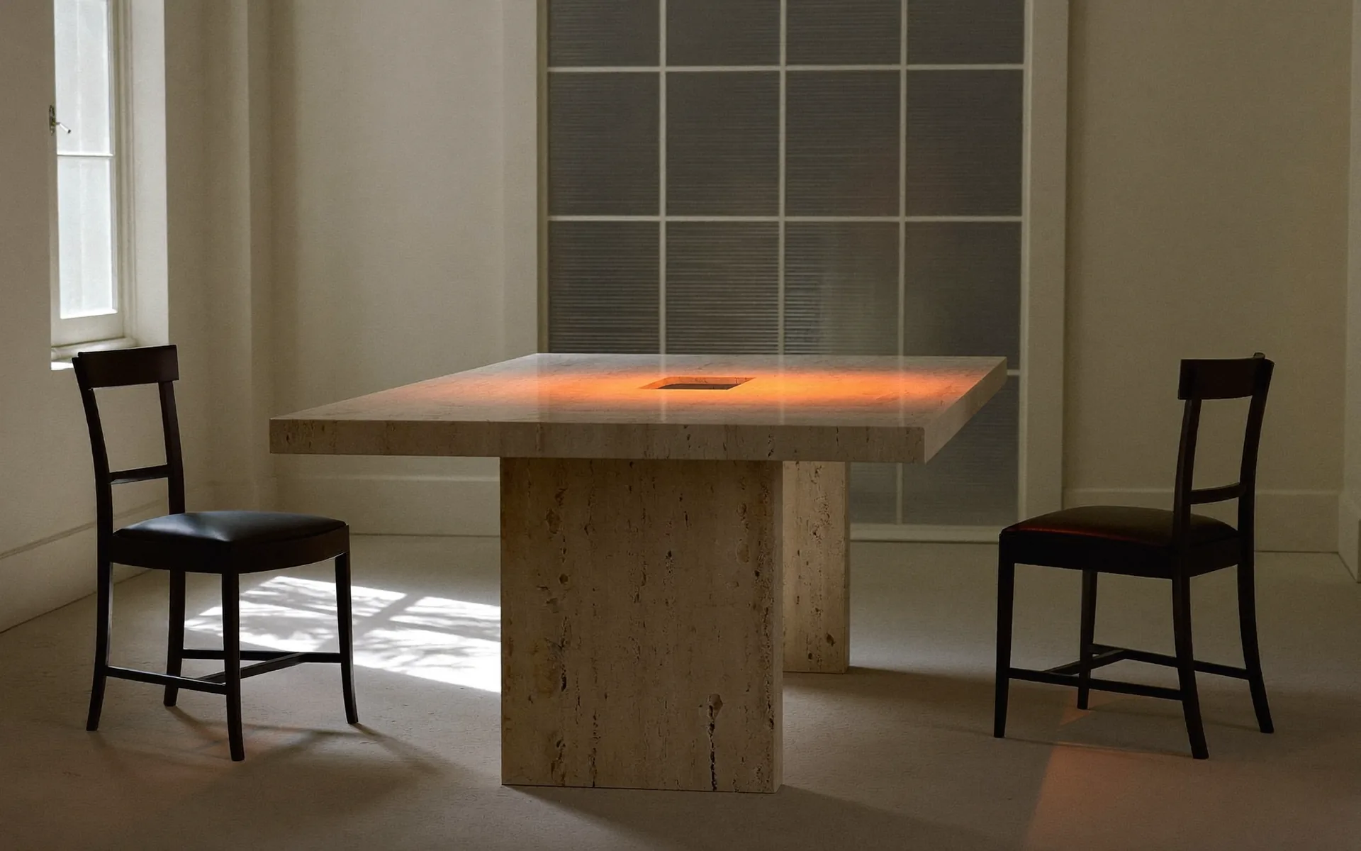 Two dark chairs facing each other across a pale travertine table, warm orange light pooling on the stone.